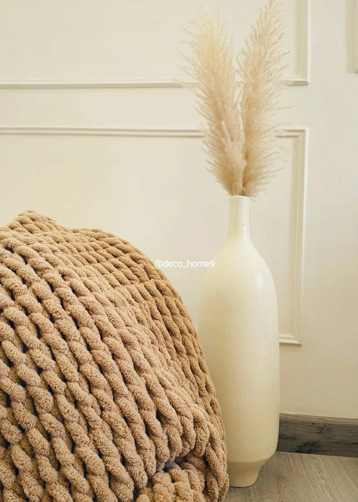 Beige textured blanket draped over a chair next to a tall white vase with dried pampas grass against a neutral wall.