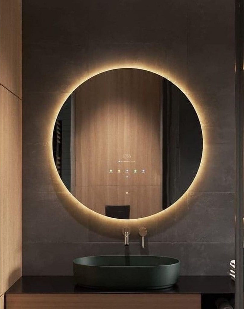Round illuminated bathroom mirror with a dark sink below on a tiled wall.