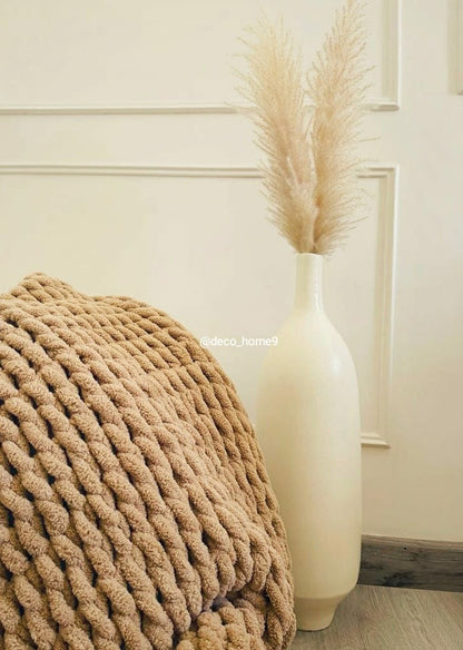 Beige textured blanket draped over a chair next to a tall white vase with dried pampas grass against a neutral wall.