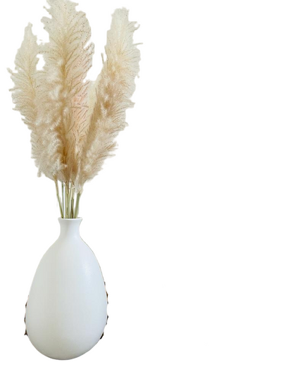 White vase with pampas grass next to a brown knitted blanket on a wooden floor.
