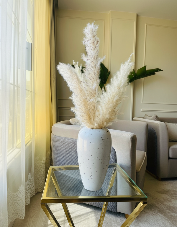 White vase with dried pampas grass on a table in a room with a chair and window.