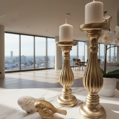 Gold candle holders with white candles on a surface, with decorative birds and a plant in the background.