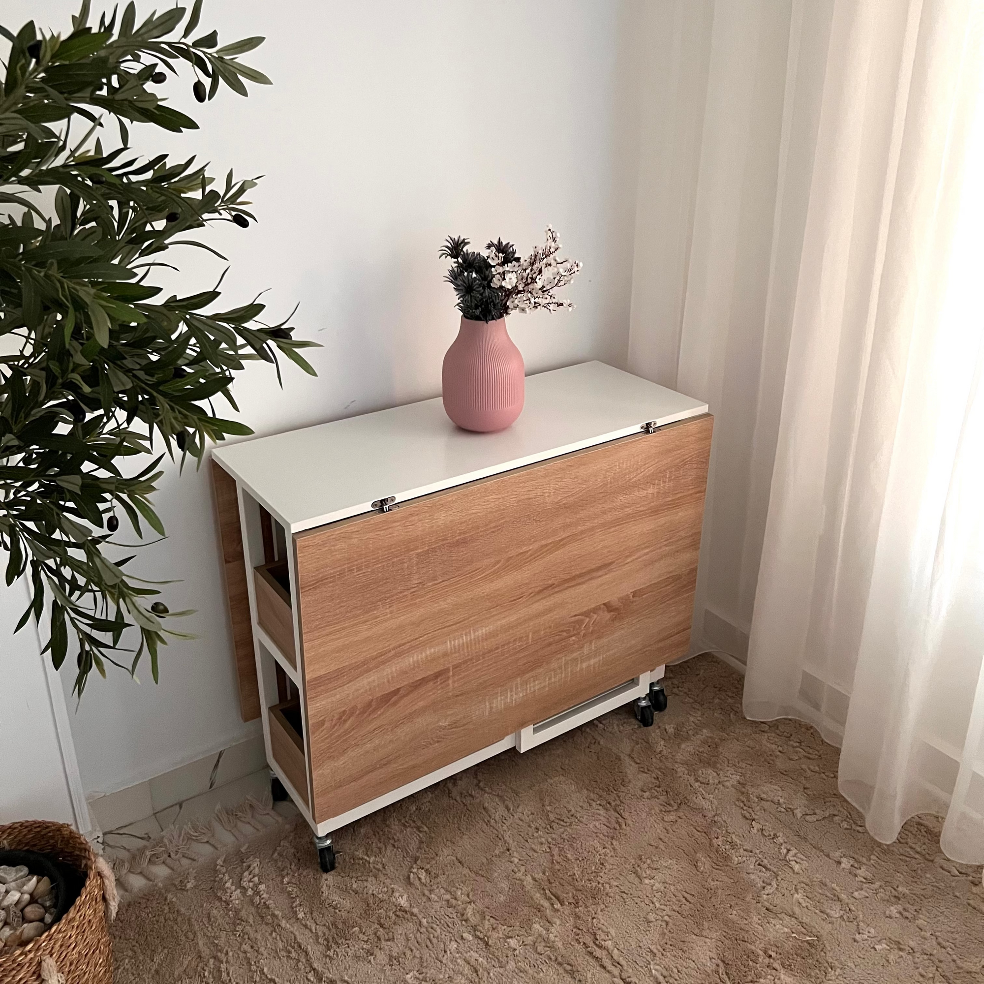 Wooden sideboard with a pink vase and plant in a room with white walls and curtains.